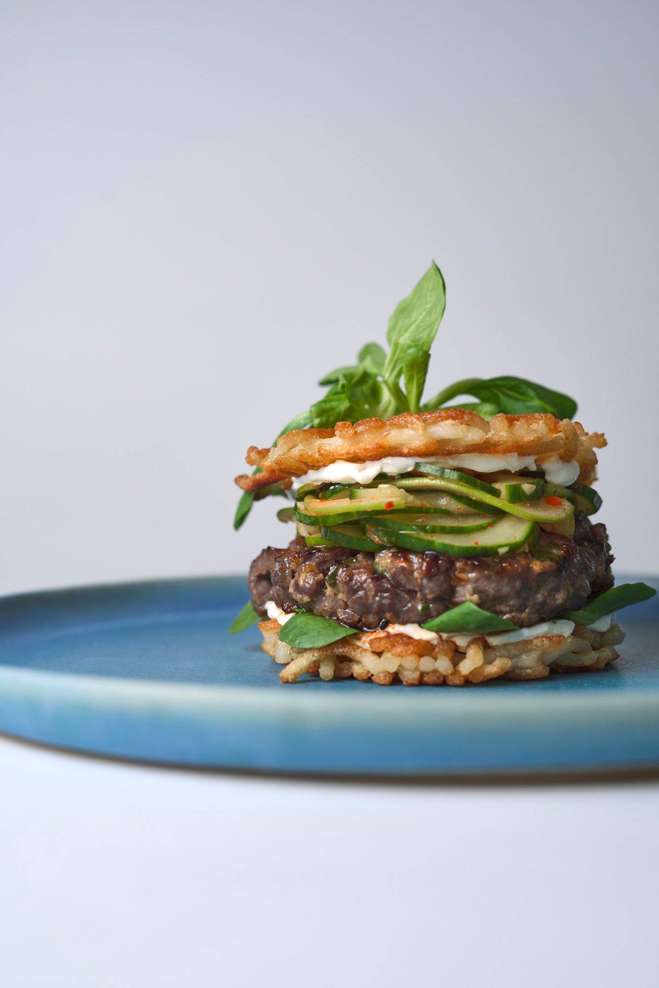 bulgogi-beef-burger-with-cucumber-kimchi bulgogi beef burger with cucumber kimchi and udon noodle buns on a blue ceramic plate with white background