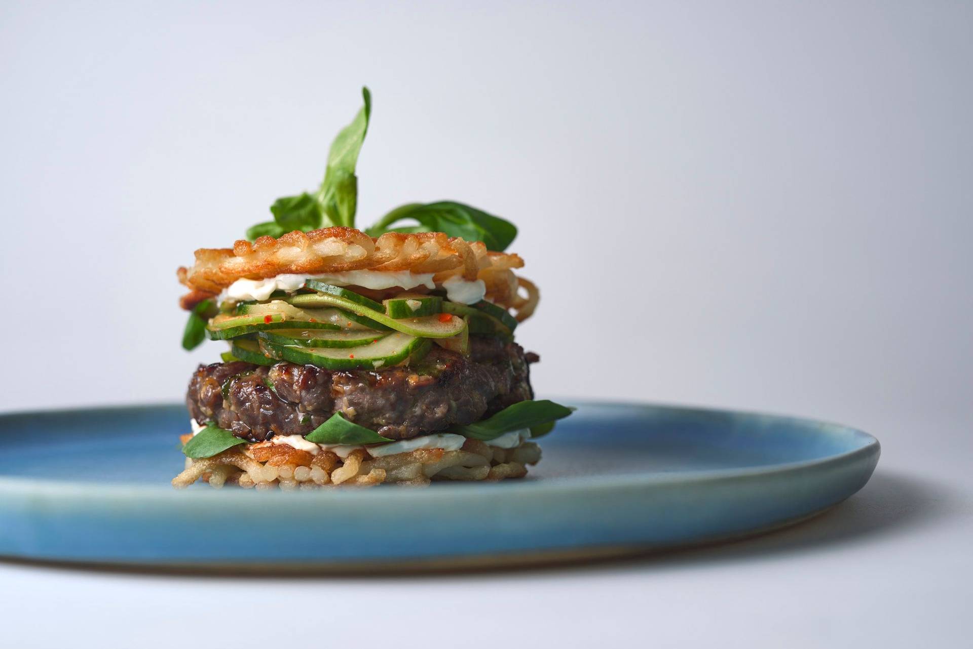bulgogi-beef-burger-with-cucumber-kimchi bulgogi beef burger with cucumber kimchi and udon noodle buns on a blue ceramic plate with white background